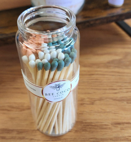 A clear glass jar without lid containing wooden matches colored in green, white, and orange, branded by Bee Coco.