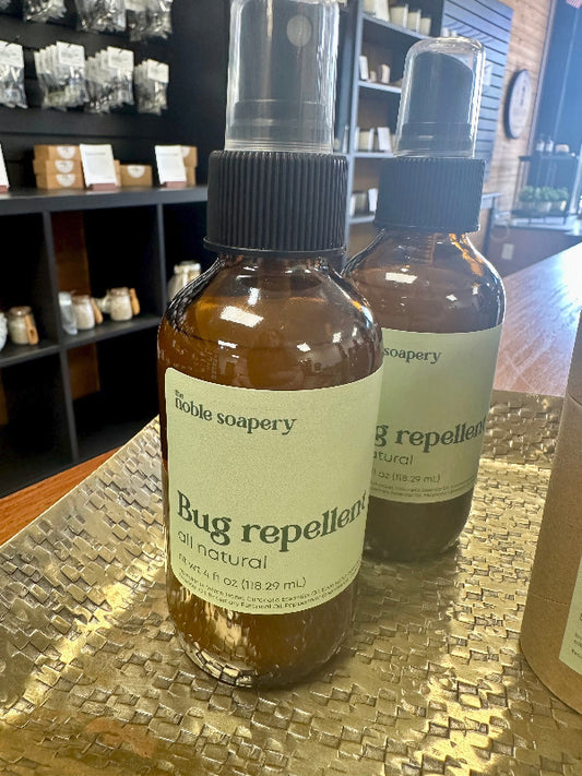 Two brown spray bottles labeled 'Bug Repellent' from The Noble Soapery on a display shelf.