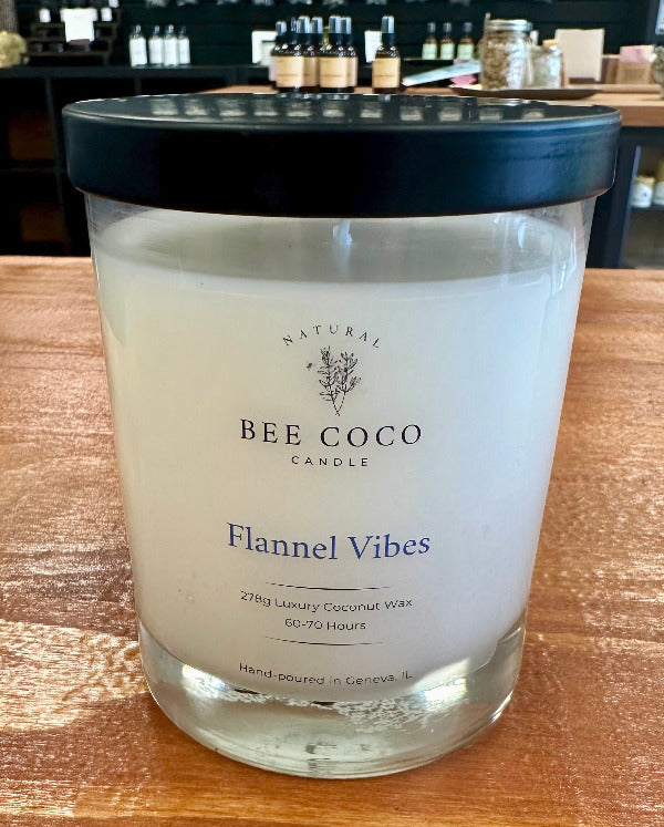 A clear glass jar candle with a black lid labeled 'BEE COCO Candle Flannel Vibes' with text indicating the scent and other attributes.