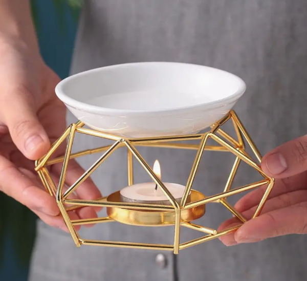 A gold-colored metal wax warmer with a geometric birdcage design, featuring a white ceramic dish and a tealight candle being held by someone.