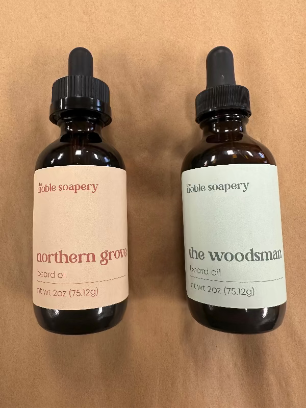Two bottles of Eagle Soapery beard oil, one labeled 'Northern Grove' and the other 'The Woodsman'. Both bottles have dropper caps and are set against a brown background.