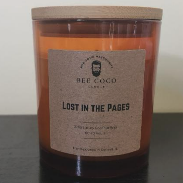 A scented candle with a brown label that reads 'BEE COCO LOST IN THE PAGES', contained in a clear glass jar with a wooden lid.