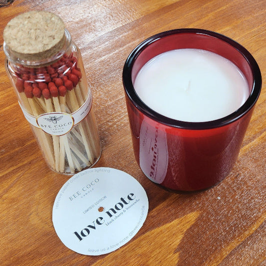 A red glass candle with a clear lid next to a container of black candle wicks and a 'Love Note' from Bee Coco Candle, placed on a wooden surface.