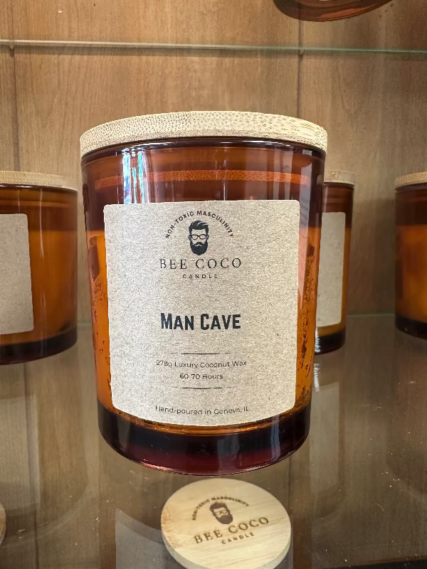 A jar candle with a label reading 'MAN CAVE' from Bee Coco. The candle is placed on a wooden surface, and there are other candles and items in the background.