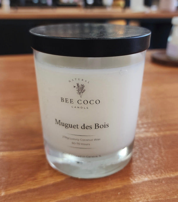 A white candle in a clear glass jar with a black lid, labeled 'Muguet des Bois' by Bee Coco Candle.