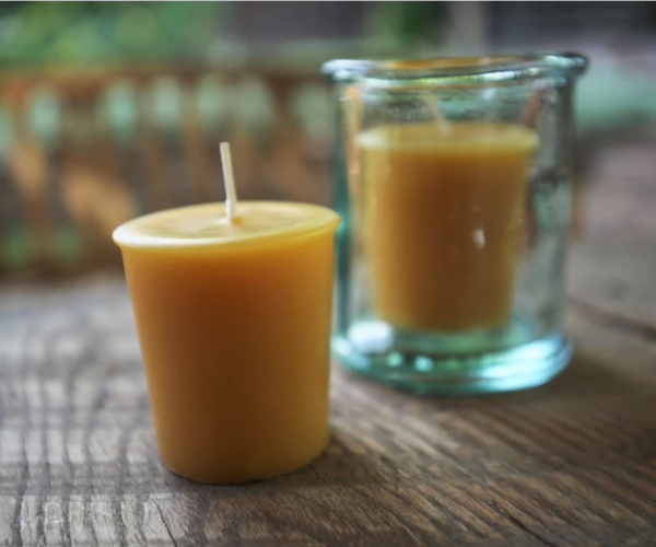 A pure beeswax votive candle in a clear glass holder, with a wooden surface and greenery in the background.