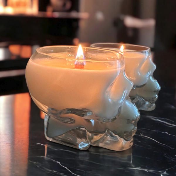 Two clear glass candles with a skull design, filled with a lighted candle, placed on a marble surface.