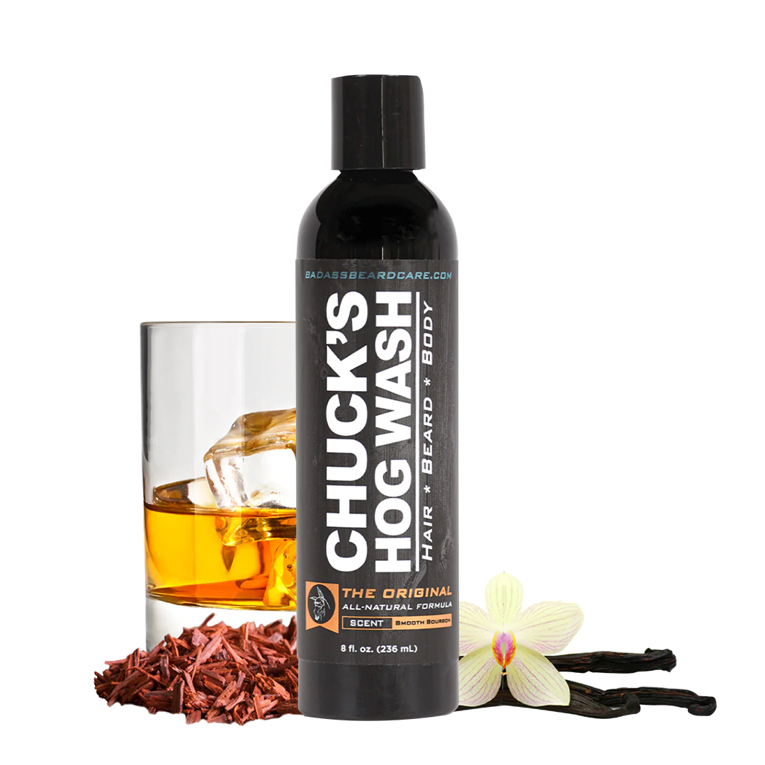 A bottle of Chuck's Hog Wash hair, beard, and body wash stands on a white background, styled with a glass of amber liquid, dried red barks, a white orchid flower, and vanilla beans. The label reads 'CHUCK'S HOG WASH' and 'HAIR BEARD BODY'.