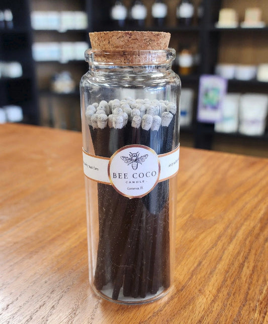 A clear glass jar with a cork lid containing wooden matches colored in black with white head, branded by Bee Coco.