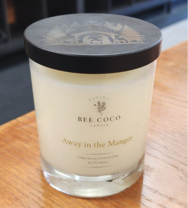 A clear glass candle jar with a black lid, labeled 'Away in the Manger' by Bee Coco Candle. The candle is placed on a wooden surface.