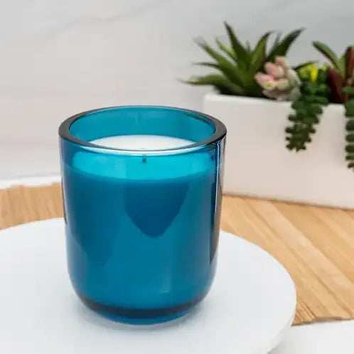 Blue candle in a glass holder on a white surface with plants in the background