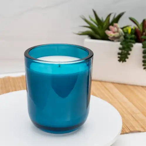 Blue candle in a glass holder on a white surface with plants in the background