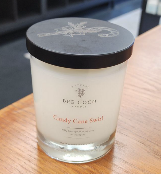 A clear candle with a black lid labeled 'Bee Coco Candle Candy Cane Swirl', placed on a wooden surface.