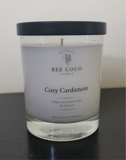 A scented candle named Cozy Cardamom by Bee Coco Candle, with a white label on a clear glass jar, black lid, and natural coconut-soy blend wax visible.