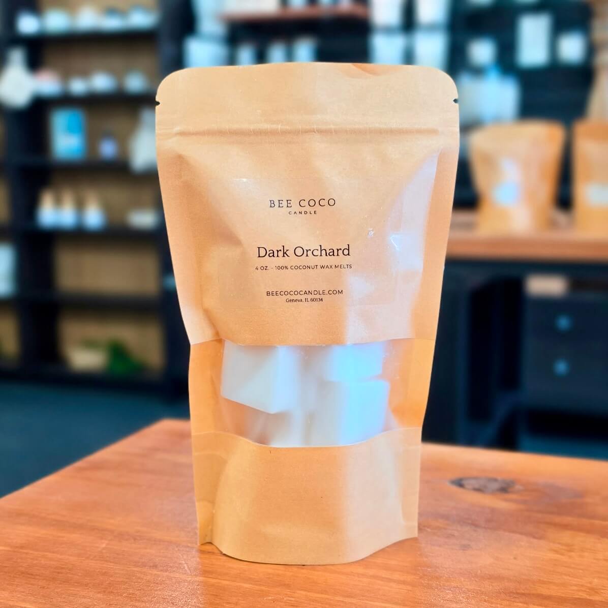 A stand-up kraft paper pouch of Bee Coco Candle Dark Orchard wax melts sits on a wooden table in a softly blurred shop interior.
