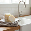 Three round dish soap refill bars of natural soap resting on a folded beige towel next to a modern kitchen sink with a stainless steel faucet.