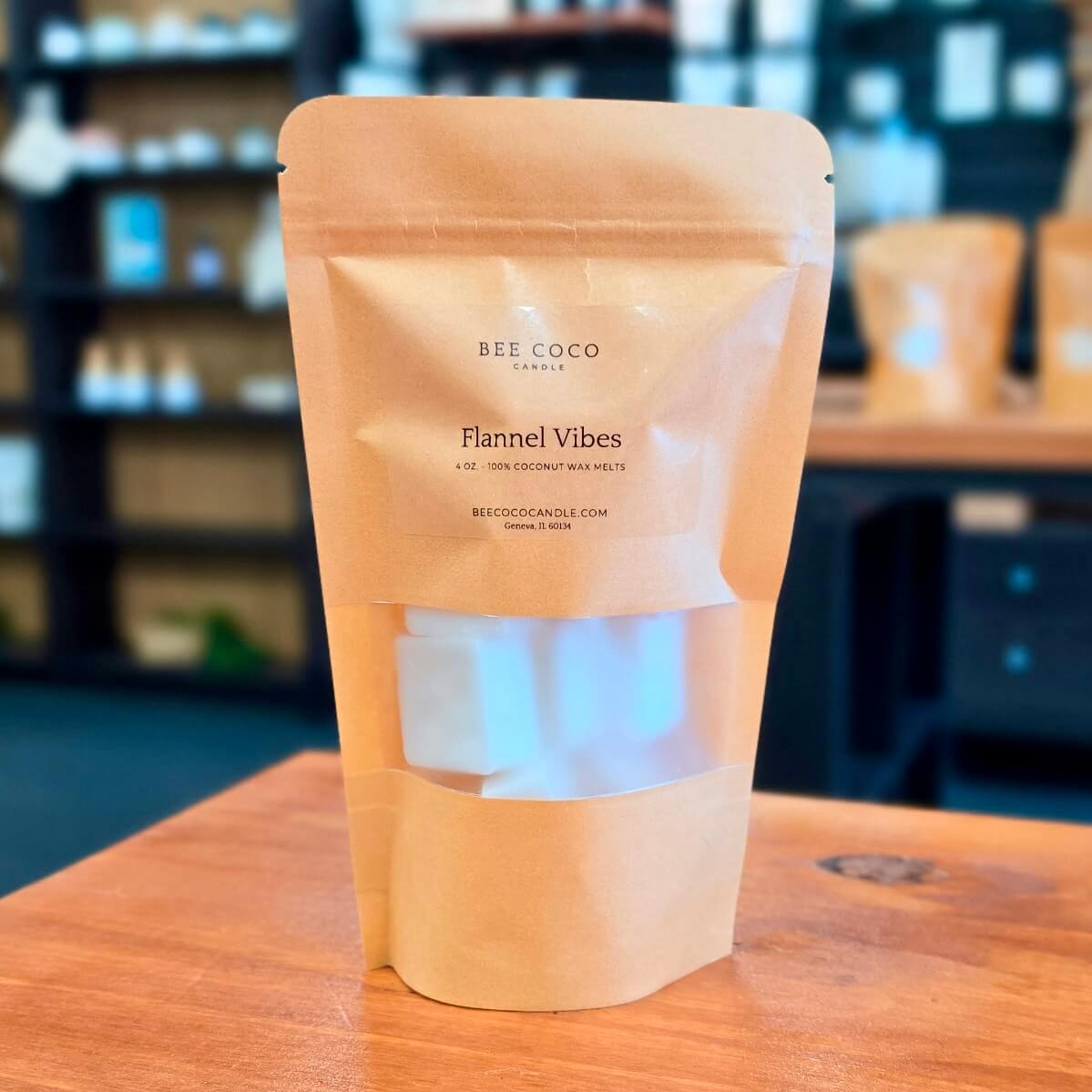 A kraft paper stand-up pouch labeled 'BEE COCO CANDLE' and 'Flannel Vibes' sits on a wooden table, with a window cutout showing white wax blocks inside. The background is a softly blurred retail or workshop setting.