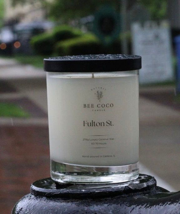 A white candle labeled 'Fulton St.' by Bee Coco in a clear glass jar with a black lid, set on a dark surface with a blurred background.