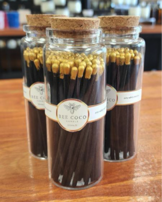 A clear glass jar with cork cap filled with gold safety matches, placed on a wooden surface.