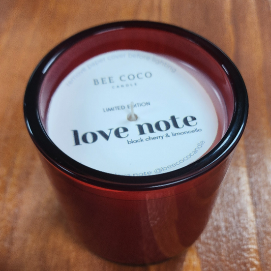 A red glass candle with a white label that reads 'love note' and indicates the fragrance notes of black cherry and lemon.