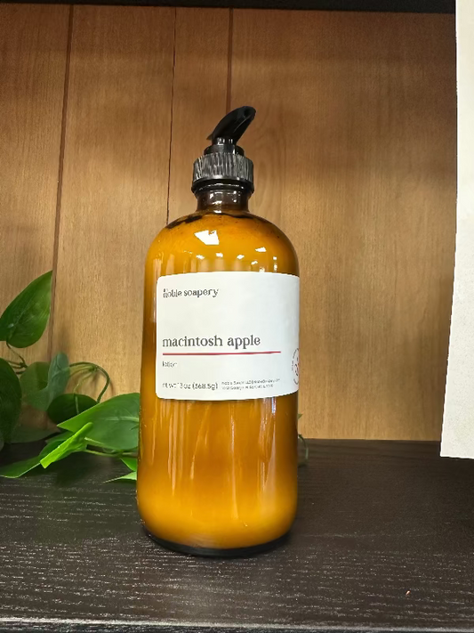A clear glass bottle labeled 'Macintosh Apple' containing a yellow liquid, placed on a wooden surface with a plant in the background.
