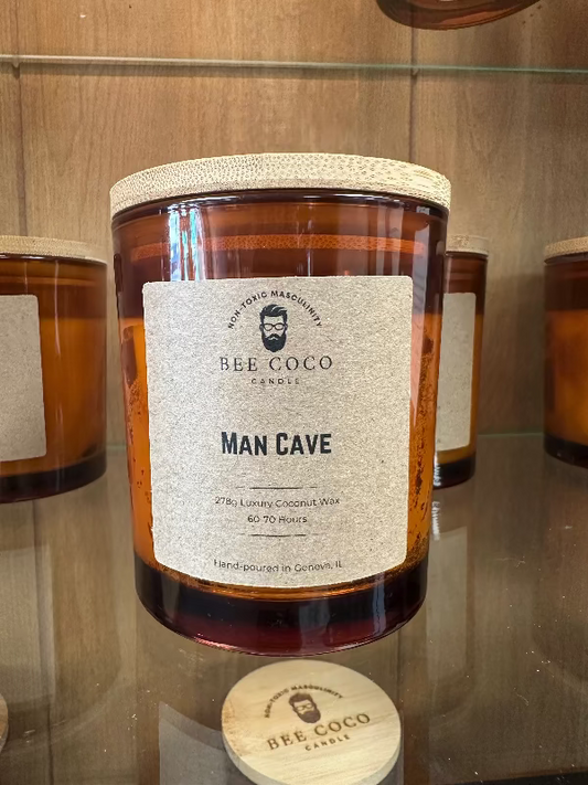 A jar candle with a label reading 'MAN CAVE' from Bee Coco. The candle is placed on a wooden surface, and there are other candles and items in the background.