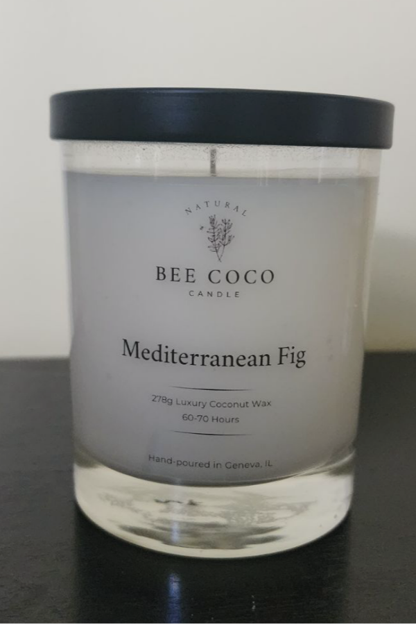 A clear glass candle jar with a black lid, labeled 'Mediterranean Fig' from Bee Coco Candle, containing a white candle, on a dark surface.