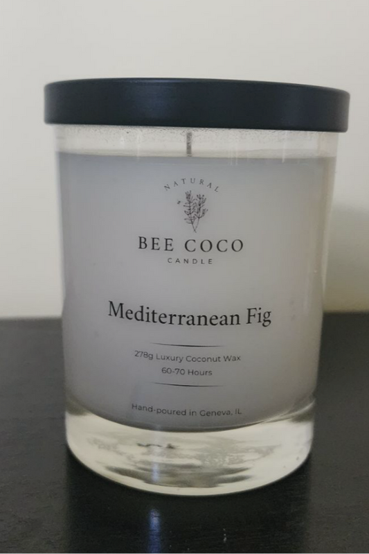 A clear glass candle jar with a black lid, labeled 'Mediterranean Fig' from Bee Coco Candle, containing a white candle, on a dark surface.