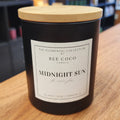 Candle labeled 'Midnight Sun' by Bee Coco Candle on a wooden surface with blurred background