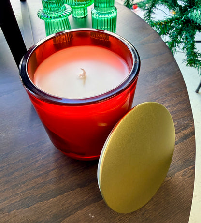 Candle in a red glass holder with a gold lid on a wooden surface, with green candle holders and a Christmas tree in the background.