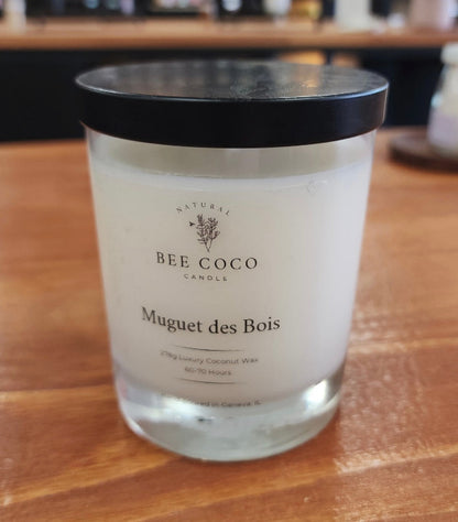 A white candle in a clear glass jar with a black lid, labeled 'Muguet des Bois' by Bee Coco Candle.
