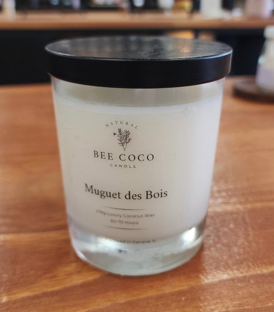 A white candle in a clear glass jar with a black lid, labeled 'Muguet des Bois' by Bee Coco Candle.