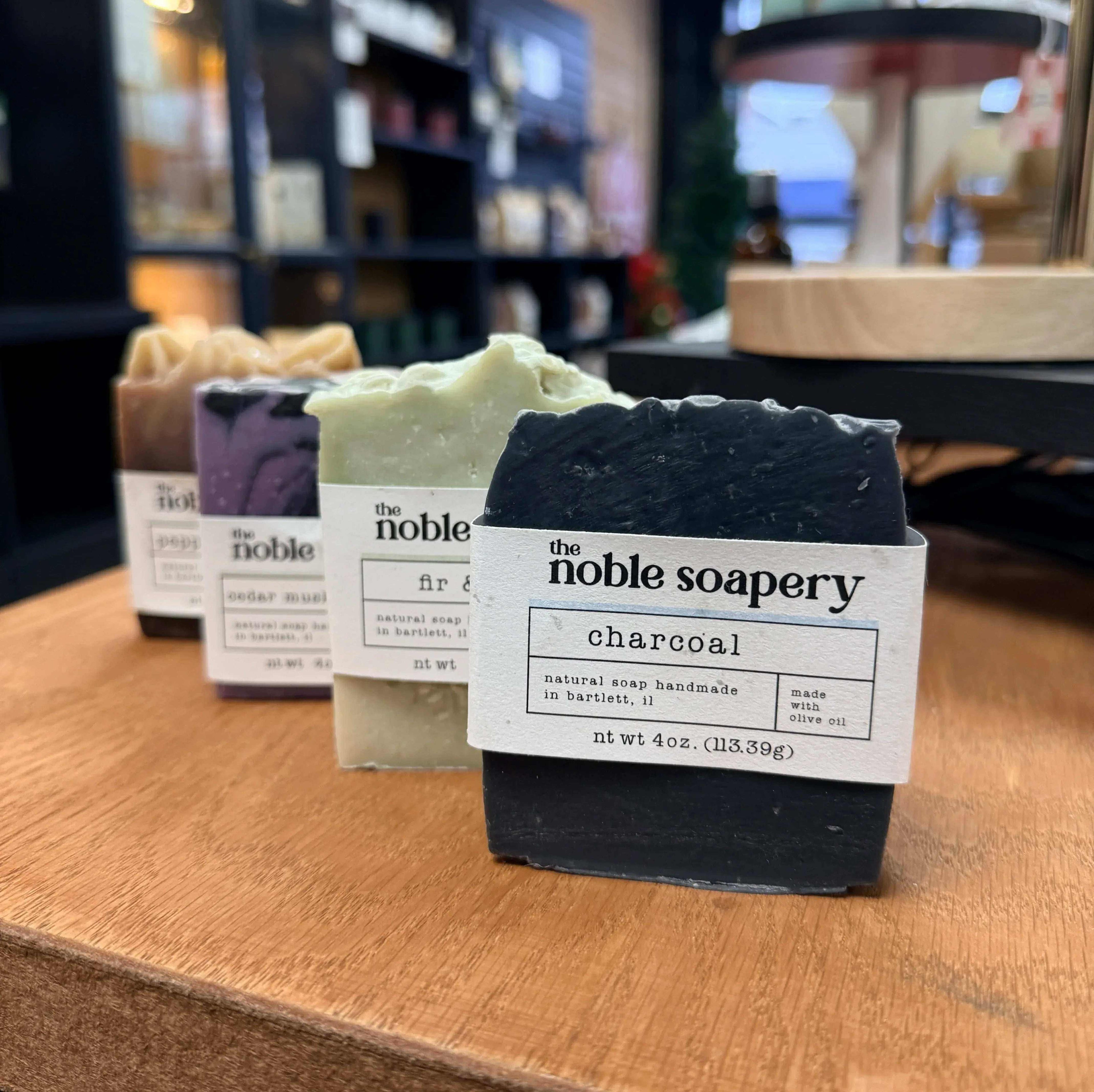 Four bars of soap, one black and one white, with 'The Noble Soapery' packaging on a wooden surface.