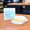A round bar of pale cream-colored solid conditioner sits on a white scrubber next to its white and light blue packaging box, which reads 'the noble soapery grapefruit & mint conditioner'. The scene is set on a wooden table with a blurred kitchen or workshop background.