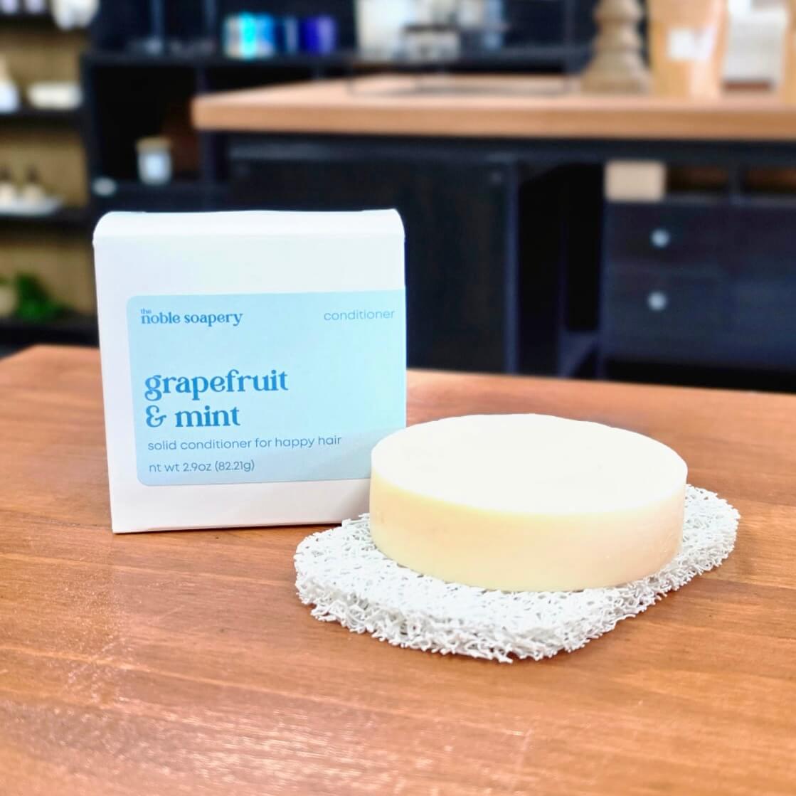 A round bar of pale cream-colored solid conditioner sits on a white scrubber next to its white and light blue packaging box, which reads 'the noble soapery grapefruit & mint conditioner'. The scene is set on a wooden table with a blurred kitchen or workshop background.