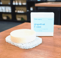 A round bar of soap sits on a textured dishcloth on a wooden table, with its white and light blue box labeled 'Bible soapery grapefruit & mint shampoo' in the blurred background.