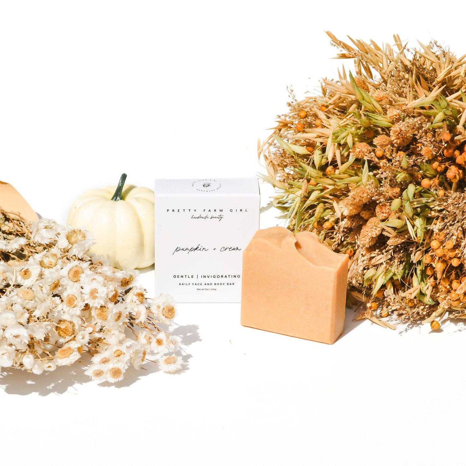 Bar of soap with a pumpkin and cream label, surrounded by dried flowers and a small pumpkin on a white background.