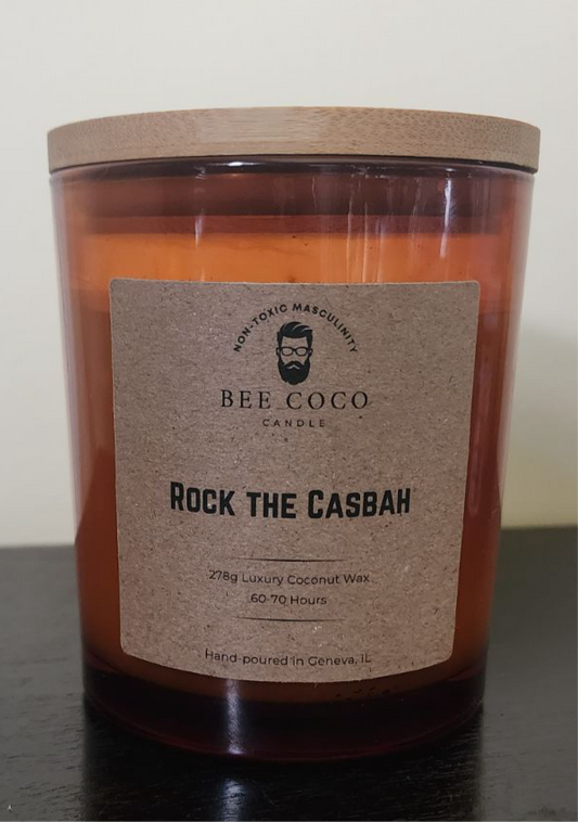 A scented candle named 'ROCK THE CASBAH' from Bee Coco Candle, with a brownish tinted glass jar and a wooden lid, featuring a label with the product name and brand.