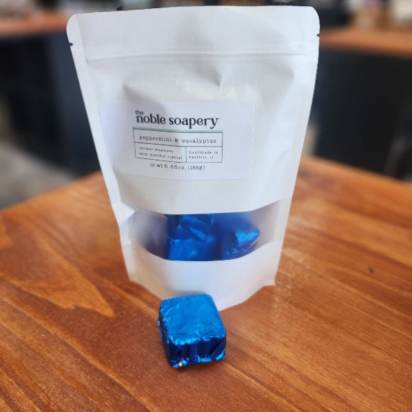 White packaging of 'The Noble Soapery' with a shower steamer wrapped in blue foil. Scent of Peppermint & Eucalyptus
