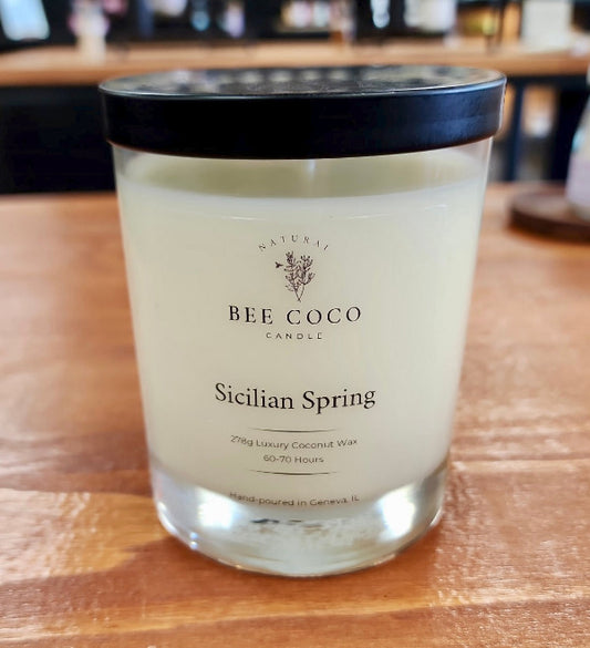 A clear glass candle jar with a black lid labeled 'Sicilian Spring' from Bee Coco. The candle is set on a wooden surface.