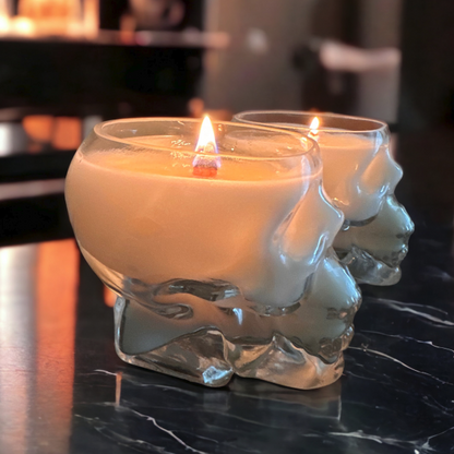 Two clear glass candles with a skull design, filled with a lighted candle, placed on a marble surface.