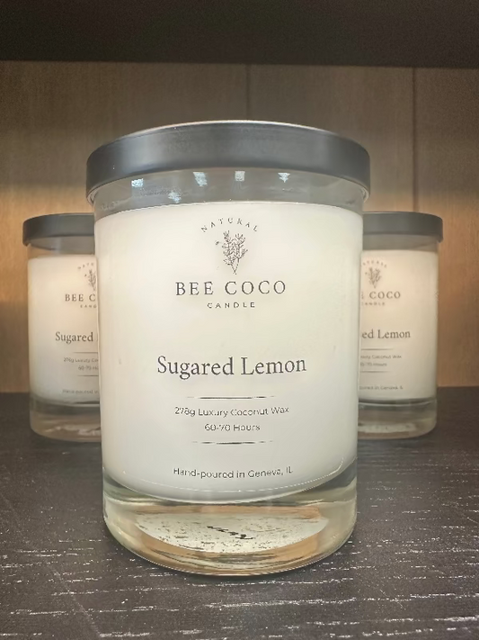 A clear glass candle jar with a label reading 'Bee Coco Sugared Lemon'. Additional jars are visible in the background.