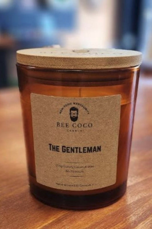 A candle named 'The Gentleman' from Bee Coco Candle, with a label on an amber glass jar, wooden lid, and a beige label with black text.