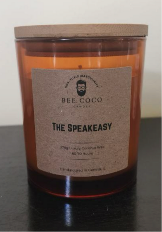 A candle jar with a label reading 'THE SPEAKEASY' from Bee Coco Candle. The jar is filled with a orange-colored wax and has a wooden lid on top.