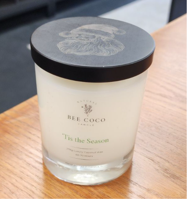 A white candle with 'Tis the Season' labeled on it, with a Santa Claus design on the lid, placed on a wooden surface.