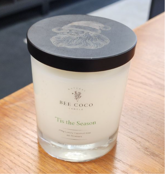 A white candle with 'Tis the Season' labeled on it, with a Santa Claus design on the lid, placed on a wooden surface.