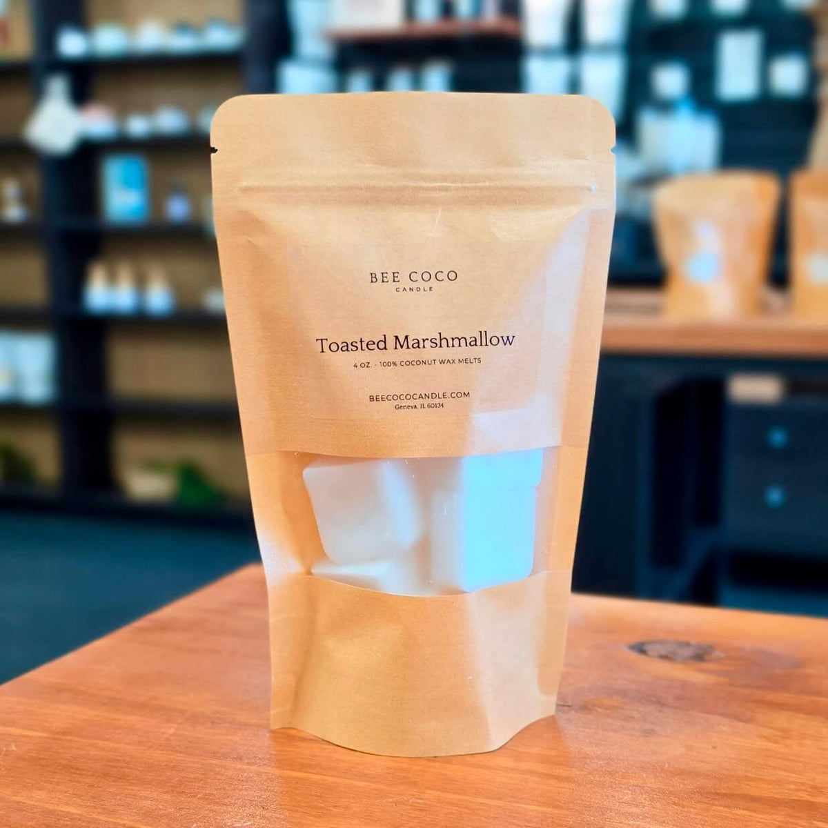 A stand-up kraft paper pouch of Bee Coco Candle in the 'Toasted Marshmallow' scent, displayed on a wooden table with a blurred shop interior in the background.