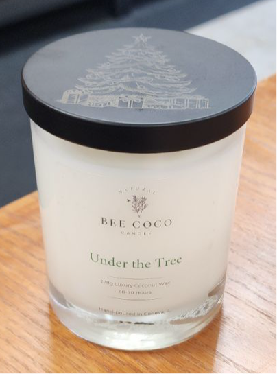 A clear glass candle with a black lid featuring a Christmas tree design, labeled 'BEE COCO UNDER THE TREE'.