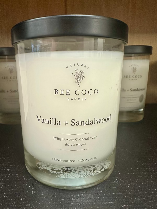 A clear glass jar candle with white wax, labeled 'Bee Coco Candle Vanilla + Sandalwood', with a black lid on top.