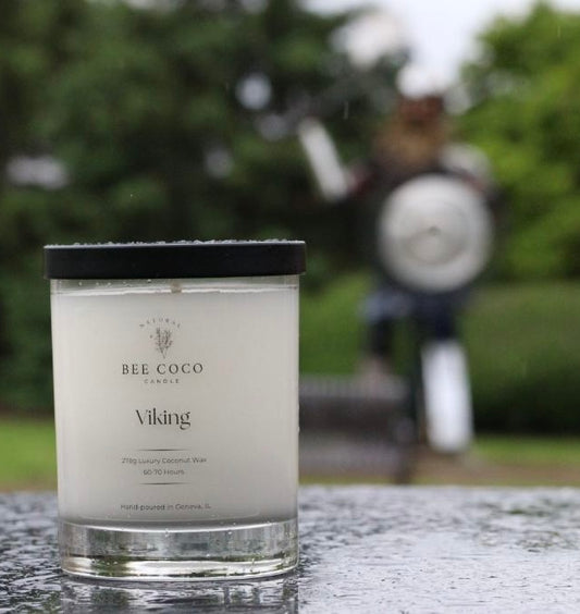 A clear glass candle jar with a black lid, labeled 'Viking', placed on a wet surface with a blurred background featuring a person playing a musical instrument.